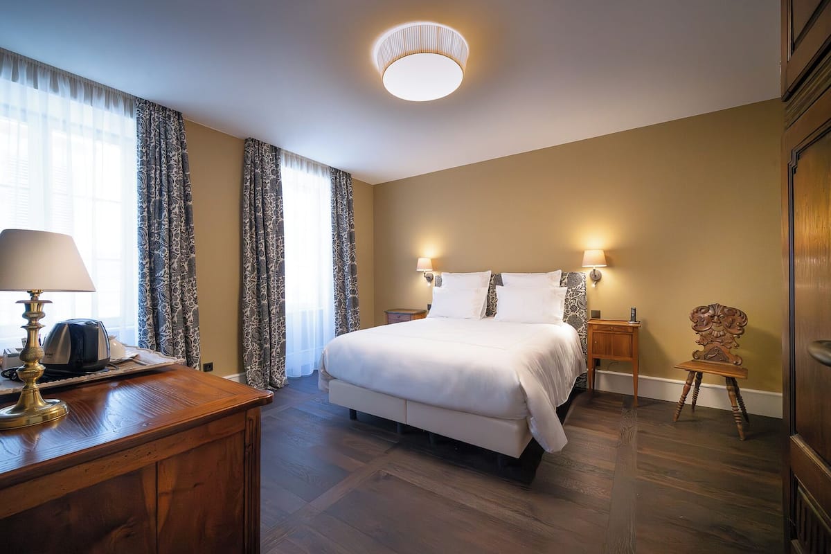 A hotel room with a bed, bedside lamps, a wooden desk with a lamp, a chair, and a ceiling light fixture.