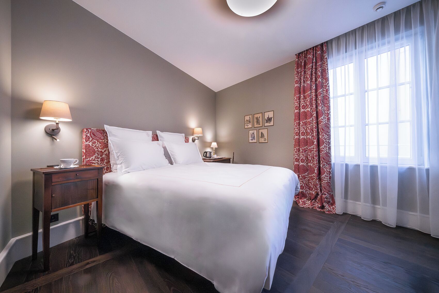 Una habitació d'hotel amb un llit gran, dues taules de nit amb làmpades, una meseta de nit, una finestra amb cortines translúcides i obra d'art penjada a la paret.