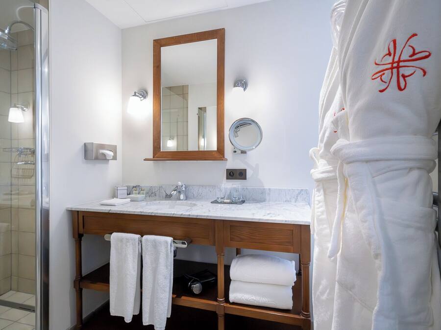 A bathroom with a glass shower enclosure, a wooden vanity with a marble countertop, a large mirror, and a robe with a red design.