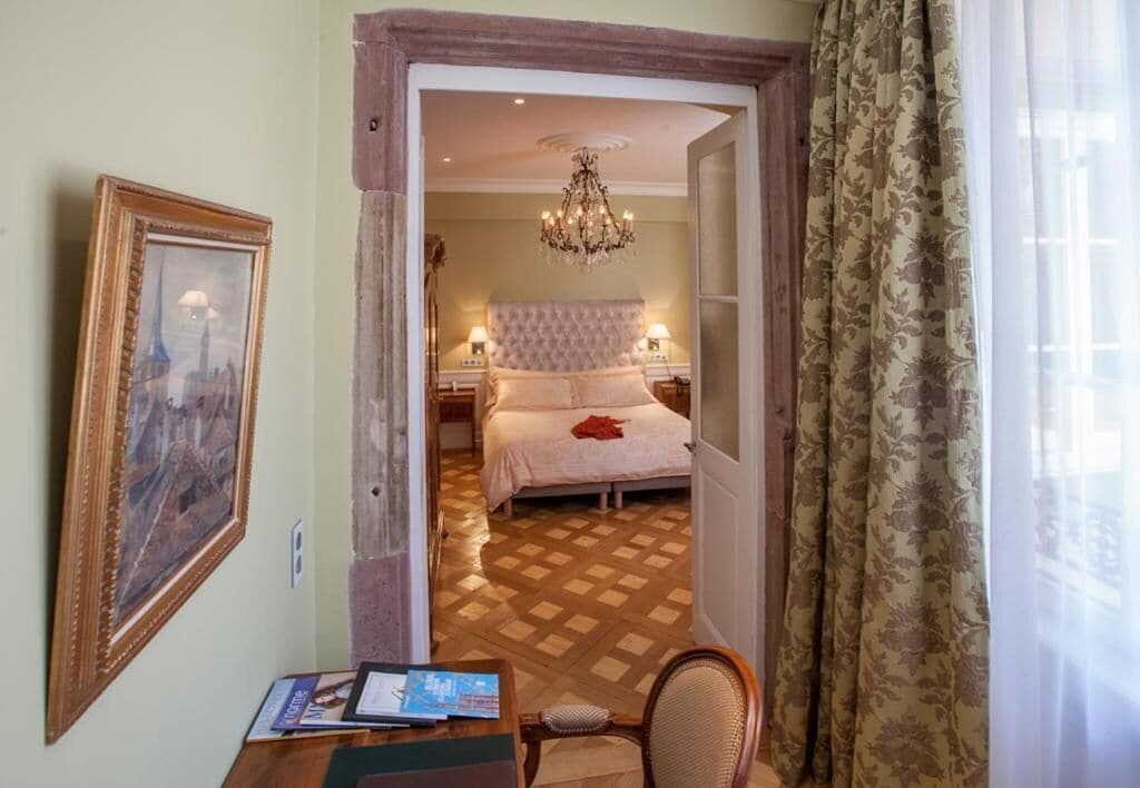 A hotel room with a bed, a chandelier, a wooden desk, and a chair.