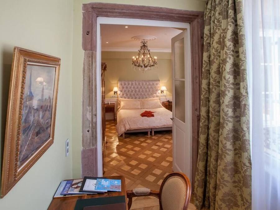 A hotel room with a bed, a chandelier, a wooden desk, and a chair.
