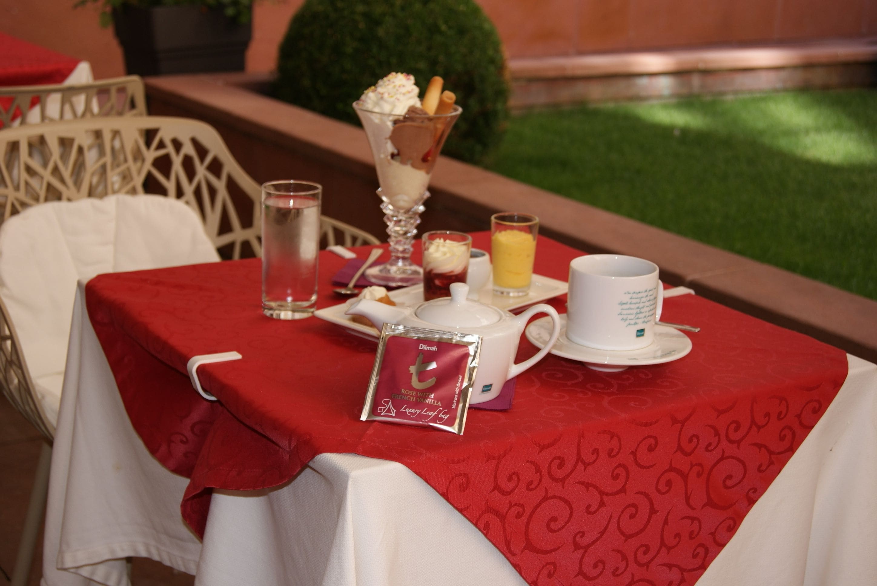 A table set for breakfast with a red tablecloth, a glass of water, a cup of coffee, a teapot, and a dessert in a glass.