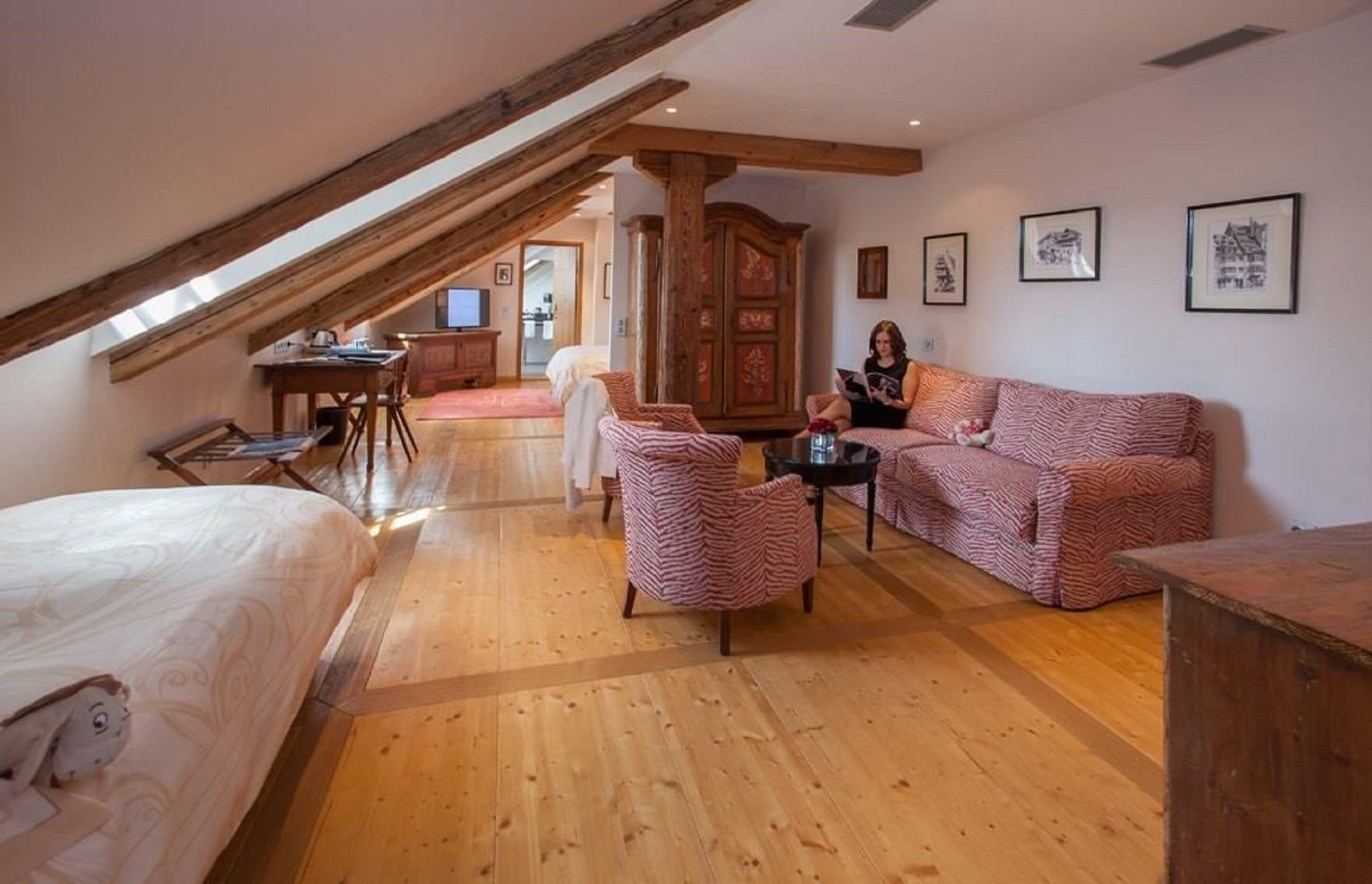 Ein gemütlicher Raum mit schrägen Wänden, Holzboden, einem Bett, einem Schlafsofa, einem Sessel, einem kleinen Tisch und einem Schrank.
