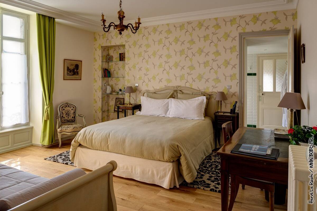 Quarto com uma cama grande, uma cadeira, uma escrivaninha, uma estante de livros e um candelabro.