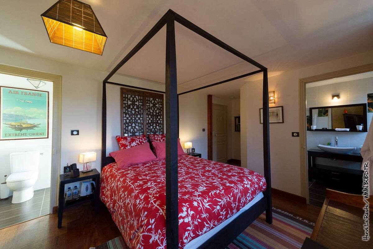 Uma cama de dossel com colcha estampada em flor vermelha, mesas de cabeceira com abajures, piso de madeira, banheiro com vaso sanitário e pia, e um pôster emoldurado na parede.