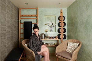 A spa room with a wooden ladder, a black mat, wicker chairs with cushions, a small table with a vase of flowers, and a shelf with toiletries.