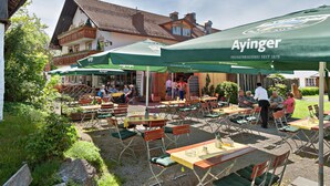 Un café en plein air avec des tables et des chaises sous des parasols verts, un bâtiment avec un toit en tuiles en arrière-plan, et des gens qui déjeunent et se promènent.