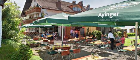 An outdoor café with tables and chairs under green umbrellas, a building with a tiled roof in the background, and people dining and walking around.