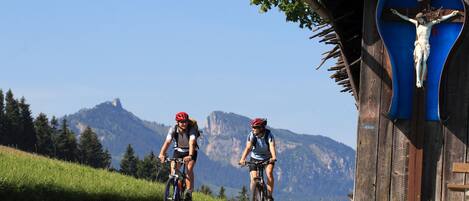 Dos ciclistas en un sendero cubierto de hierba, con una estructura de madera al fondo que presenta una cruz.