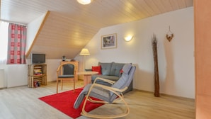 Un salon chaleureux avec un plafond en bois, un canapé gris, un fauteuil à bascule, un tapis rouge et une petite table avec une lampe.