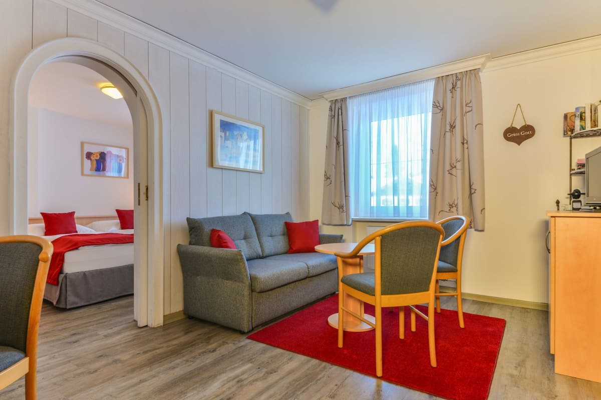 Una sala de estar moderna con un sofá gris, cojines rojos, una mesa de comedor de madera con sillas, un tapete rojo y una zona de cocina con un mueble y un letrero colgante.
