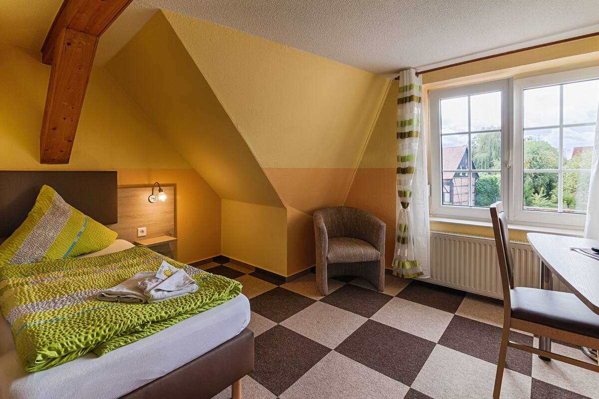 A room with a bed, a chair, a table, and a window with a view of buildings and trees.