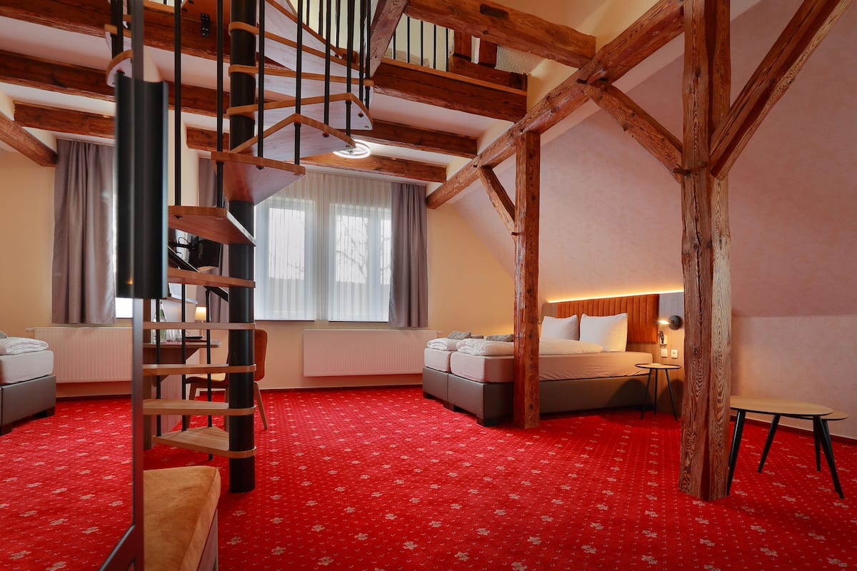 A room with a red carpet, wooden beams, a spiral staircase, a bed, a desk, and a window with curtains.