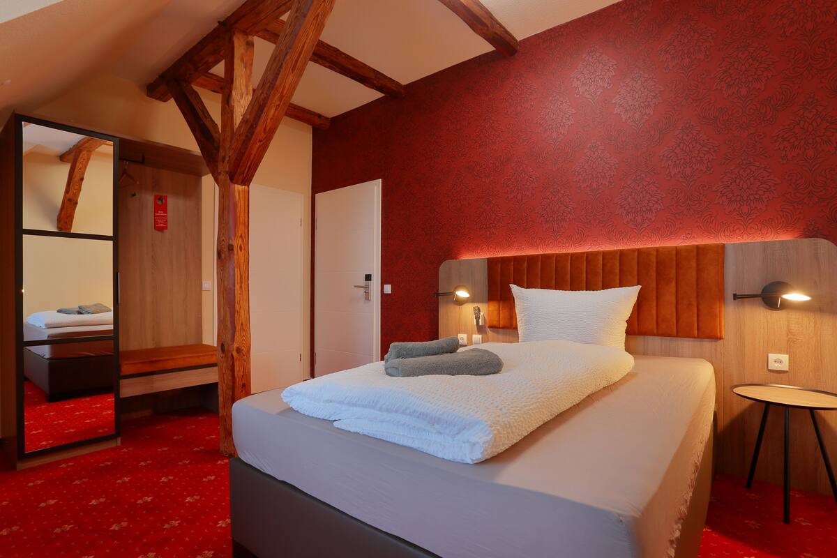 A hotel room with a bed, a wooden headboard, a red patterned wall, and a small table with a lamp.