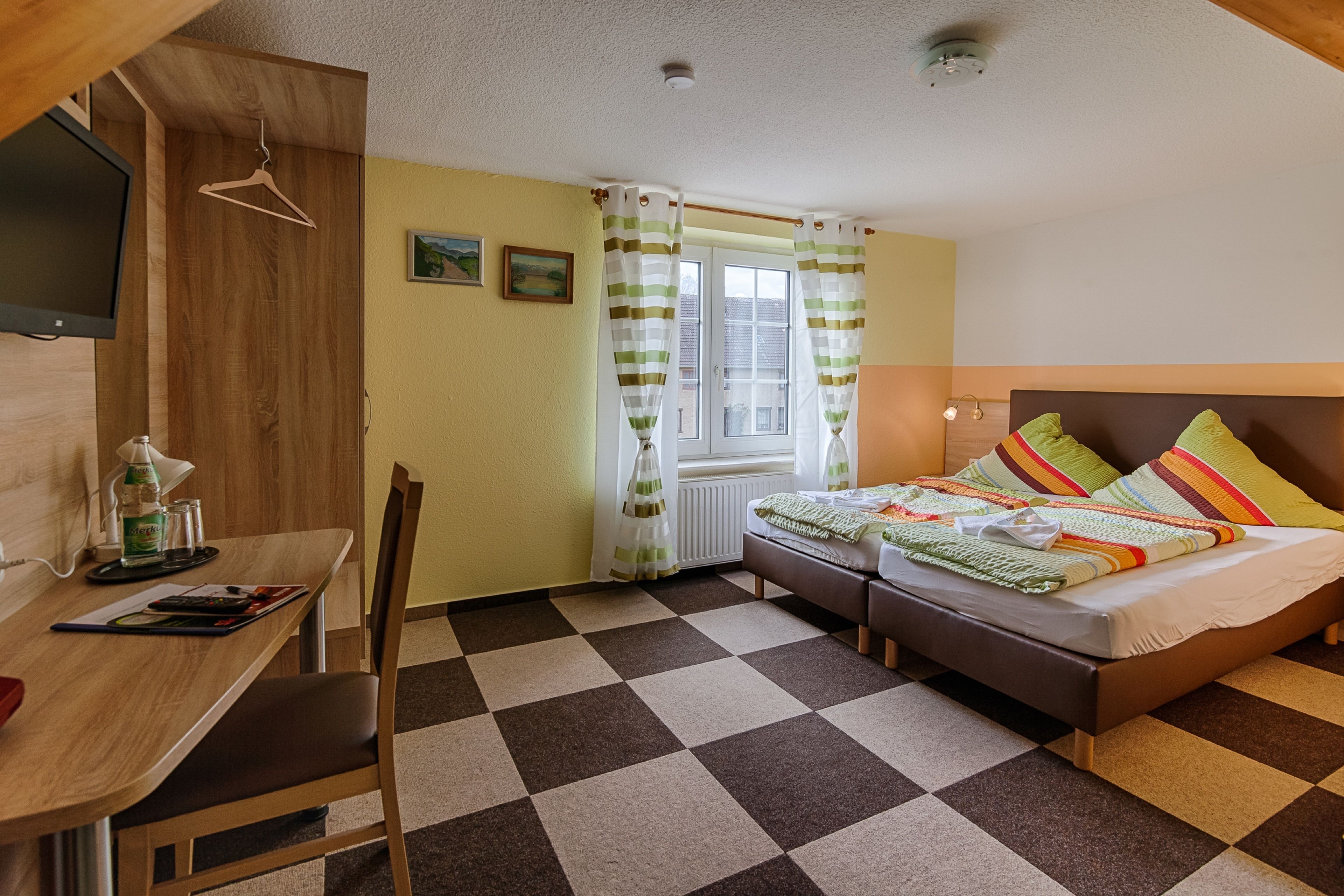 A room with a checkered floor, a bed with colorful pillows, a desk with a lamp, and a TV mounted on the wall.