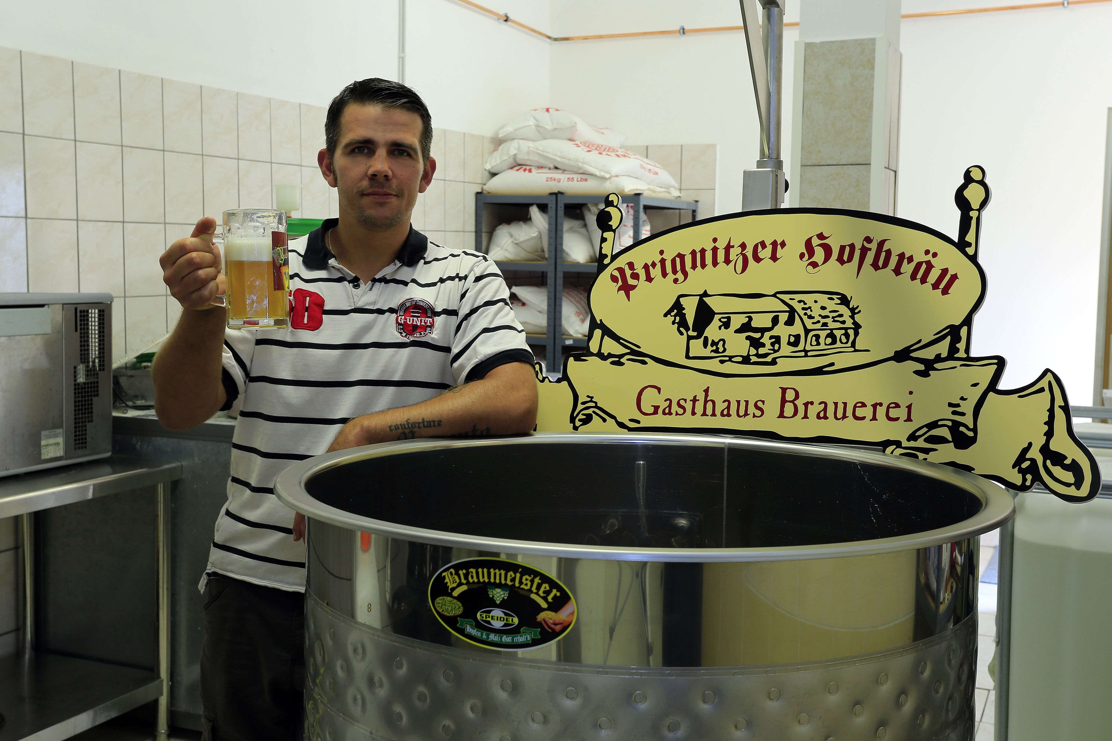 Un hombre sostiene una jarra de cerveza en una cocina con un tanque de elaboración grande y un letrero que dice 