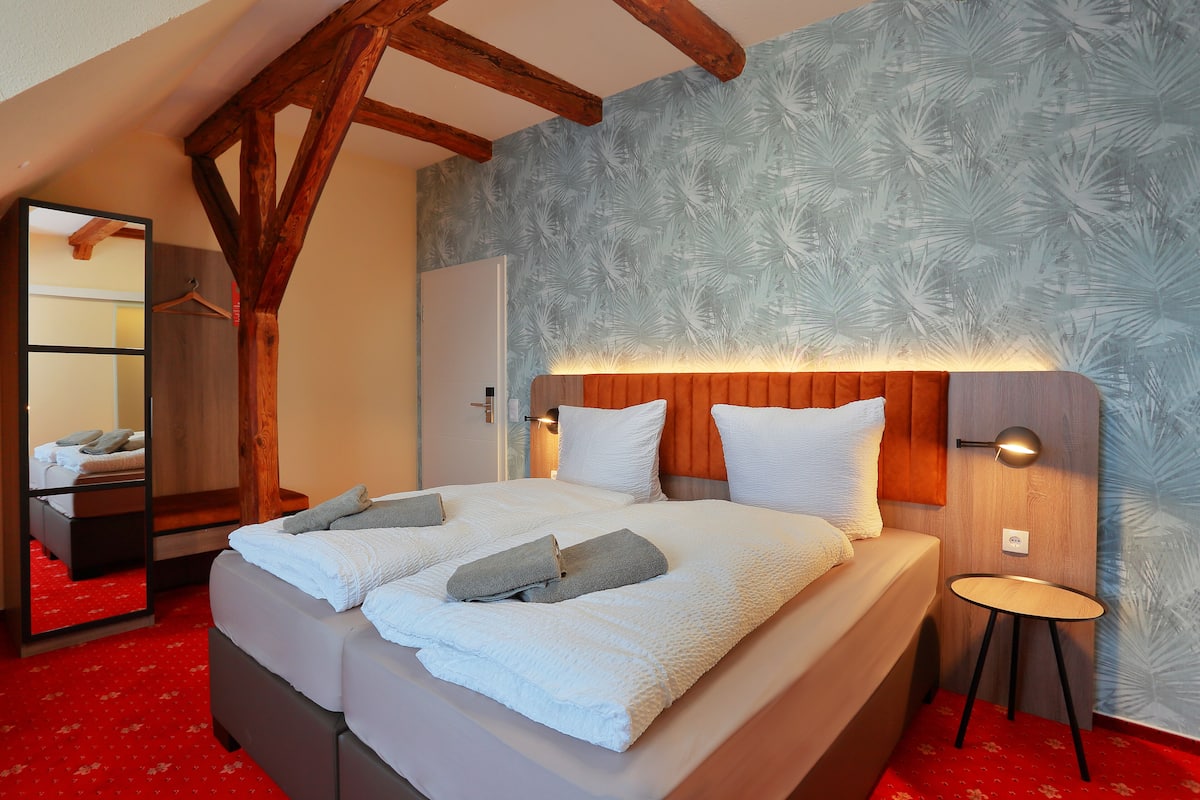 A neatly made bed with white linens and gray towels, a wooden headboard, and a small bedside table with a lamp.