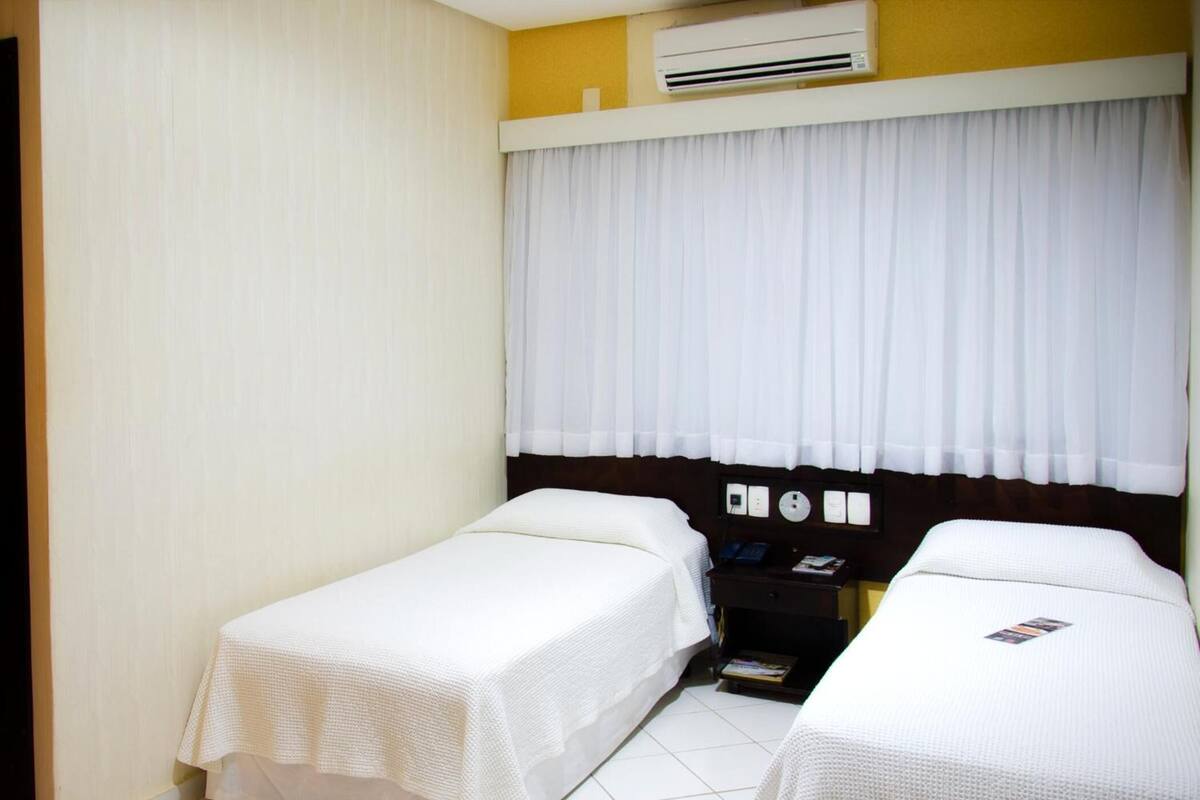 Une chambre d’hôtel avec deux lits simples, une table de chevet avec un téléphone et un livre, et un appareil de climatisation placé au-dessus de la fenêtre.