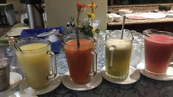 Una mesa de buffet con diversas bebidas, entre ellas zumos y batidos, y una máquina de café al fondo.