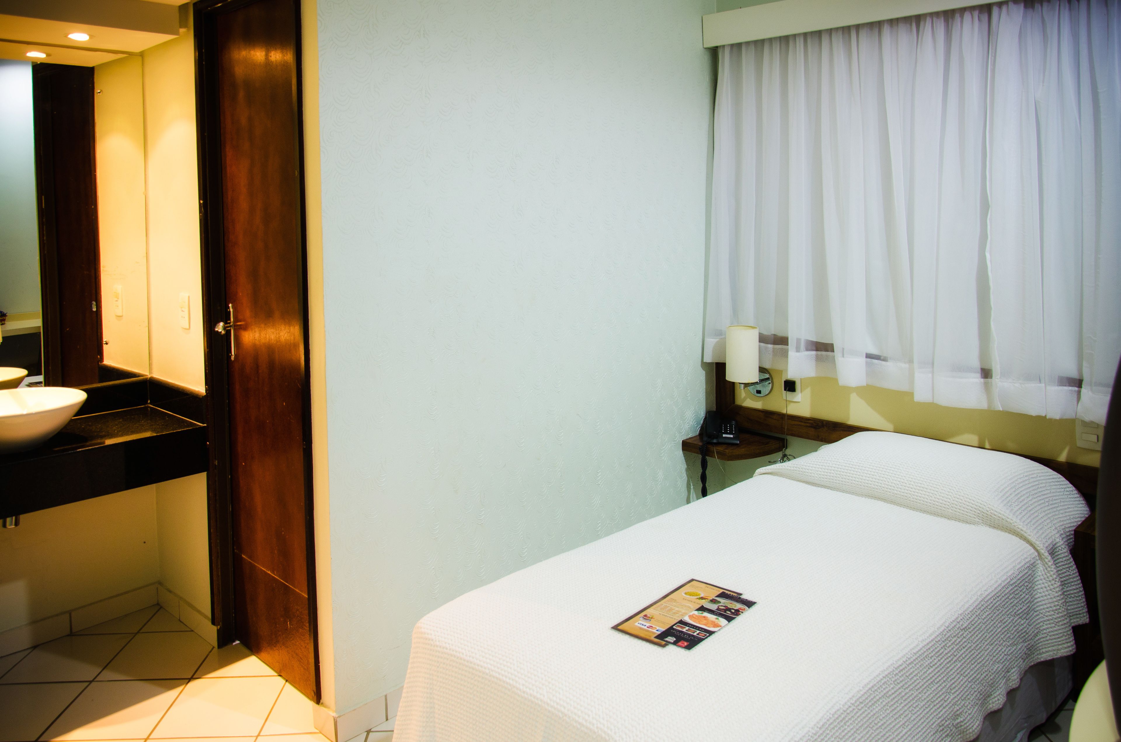 Habitación de hotel con cama, mesita de noche, lámpara, ventana con cortinas y baño con lavabo y espejo.