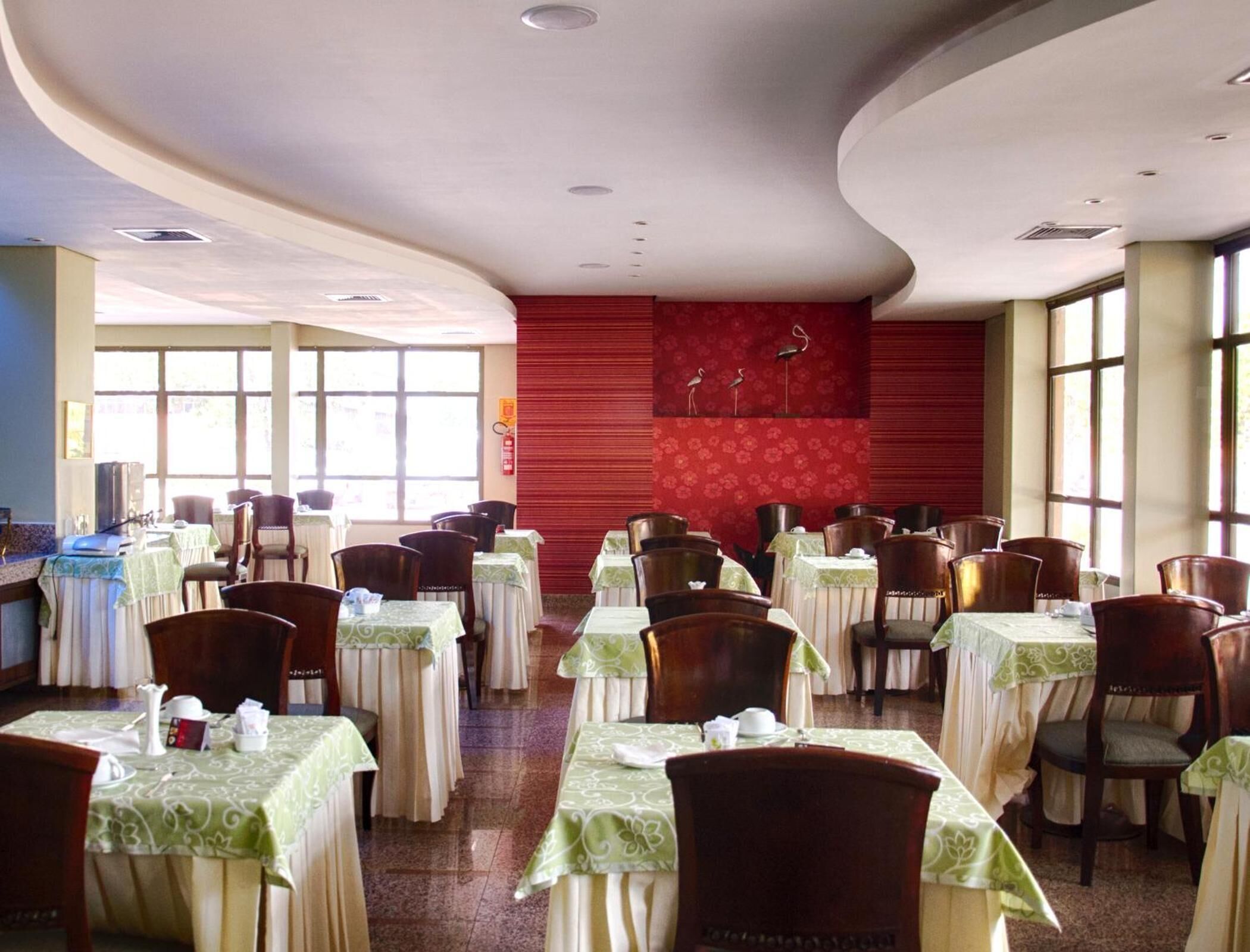 Un comedor con mesas dispuestas para el servicio, un muro rojo decorativo y amplias ventanas que dejan entrar luz natural.