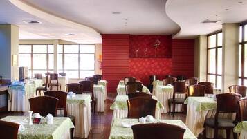 Un comedor con mesas dispuestas para el servicio, un muro rojo decorativo y amplias ventanas que dejan entrar luz natural.