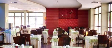 Un comedor con mesas dispuestas para el servicio, un muro rojo decorativo y amplias ventanas que dejan entrar luz natural.