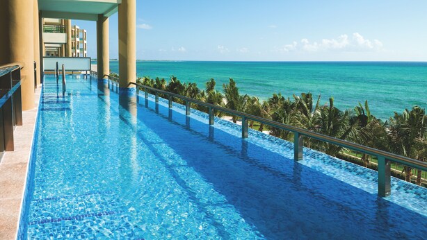 Una piscina larga y cristalina de color azul, con borde de azulejos, que da a una playa y al océano.