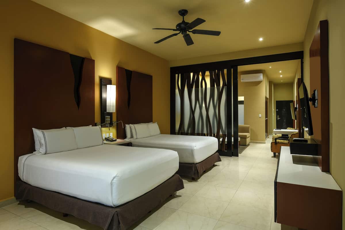 A hotel room with two beds, a desk, a chair, a TV, and a ceiling fan.