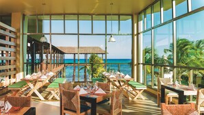 Un restaurante con mesas y sillas de madera, ventanas amplias con vistas al océano y una estructura con techo de paja.