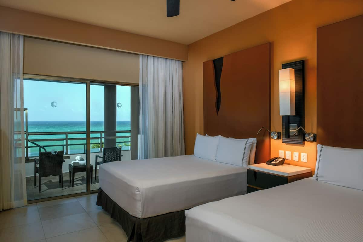 A hotel room with two beds, a balcony with a view of the ocean, and a nightstand with a lamp.