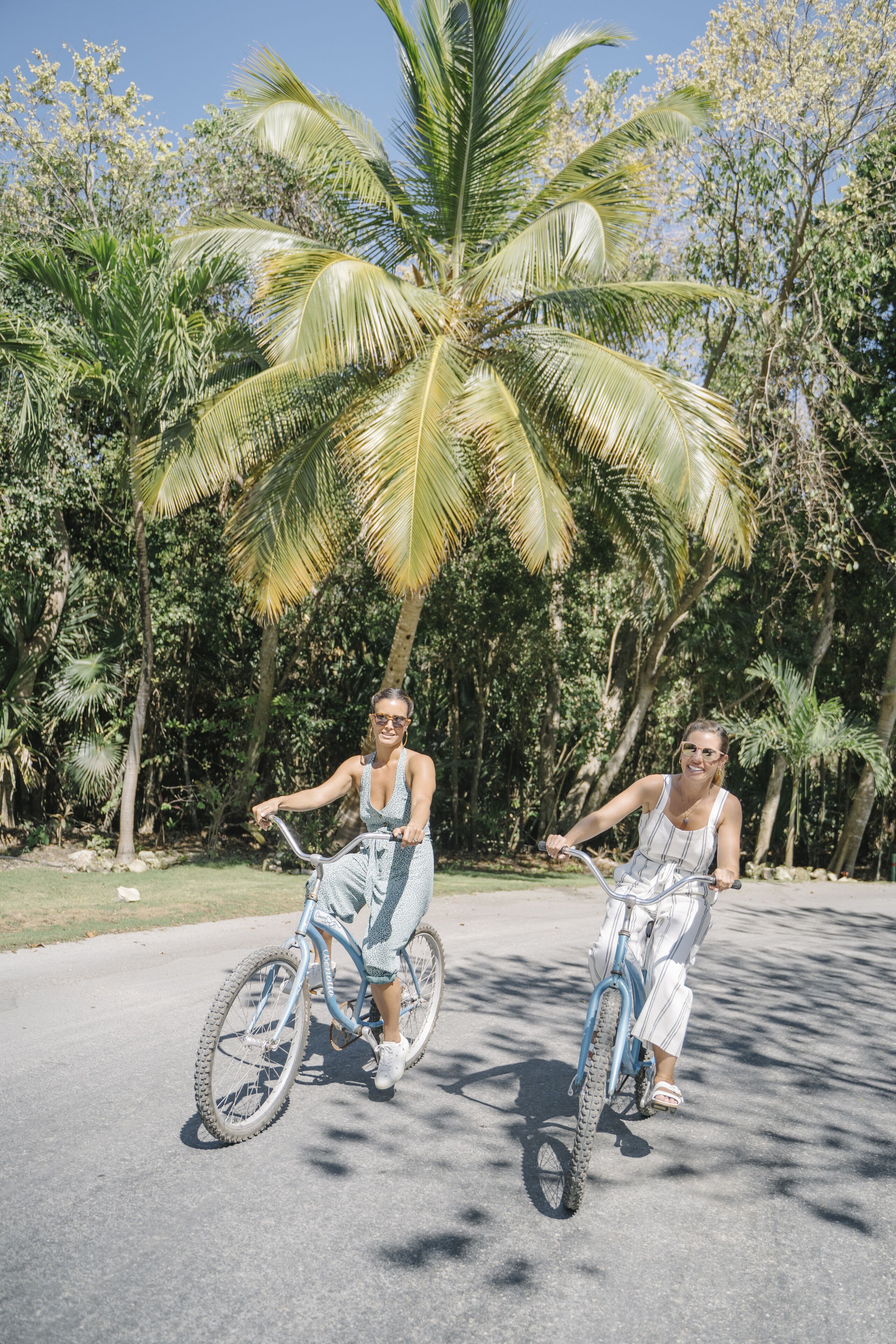 Dos personas montando en bicicleta por un sendero pavimentado, con palmeras al fondo.