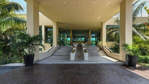 Una entrada de hotel moderna con una escalera central, rodeada de plantas en macetas y palmeras.