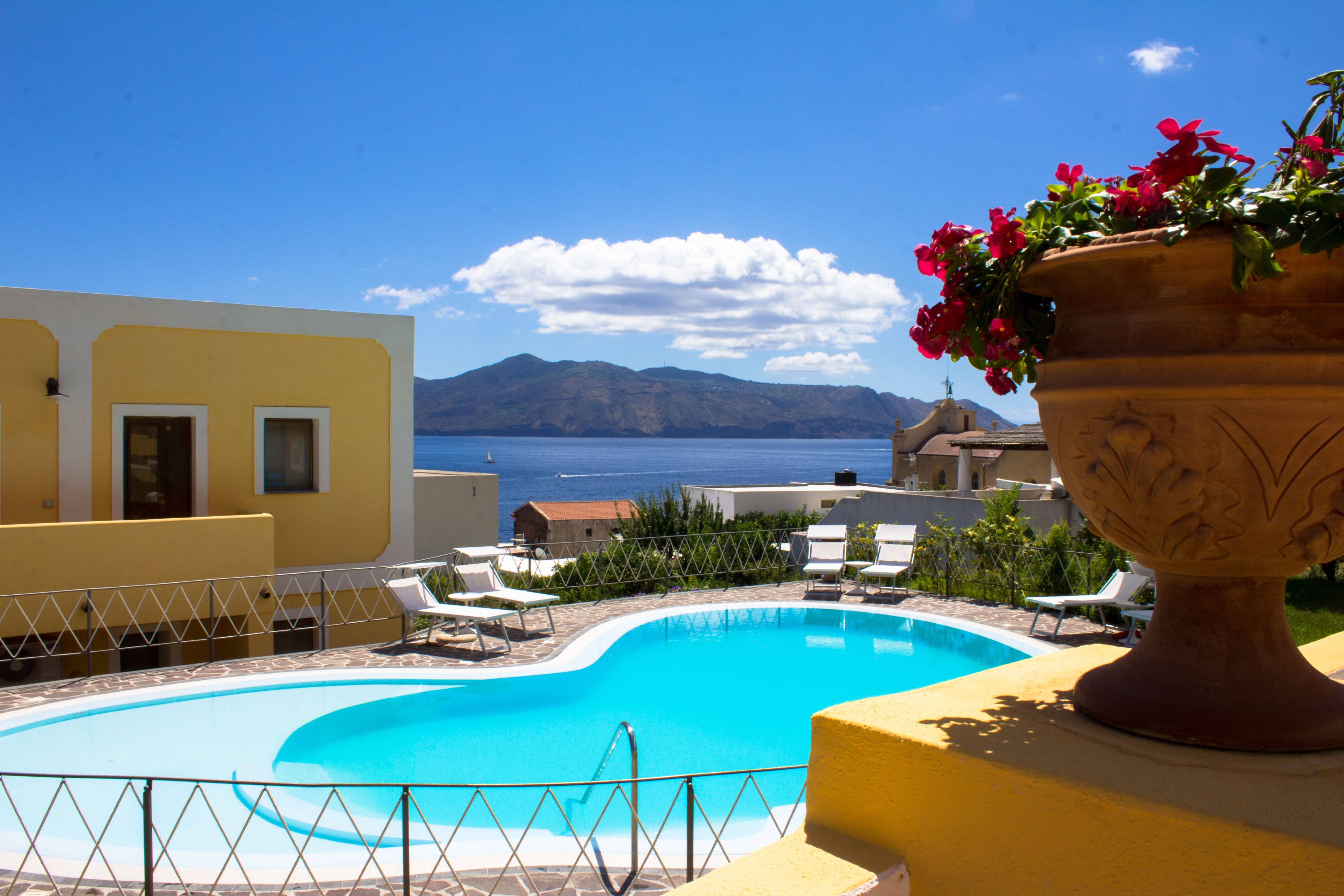 A pool area with a view of the sea and mountains, surrounded by yellow buildings and white lounge chairs.