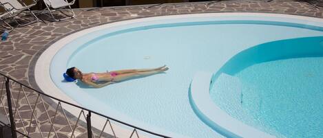 A woman in a pink bikini lying on a lounge chair by a pool with a stone-tiled deck and a metal railing.