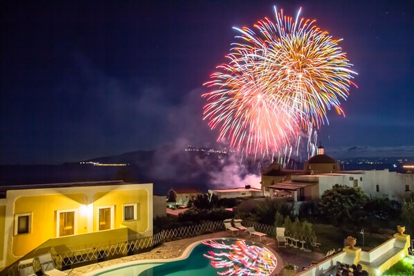 Vista nocturna de un hotel con piscina, zona de patio y fuegos artificiales a lo lejos.