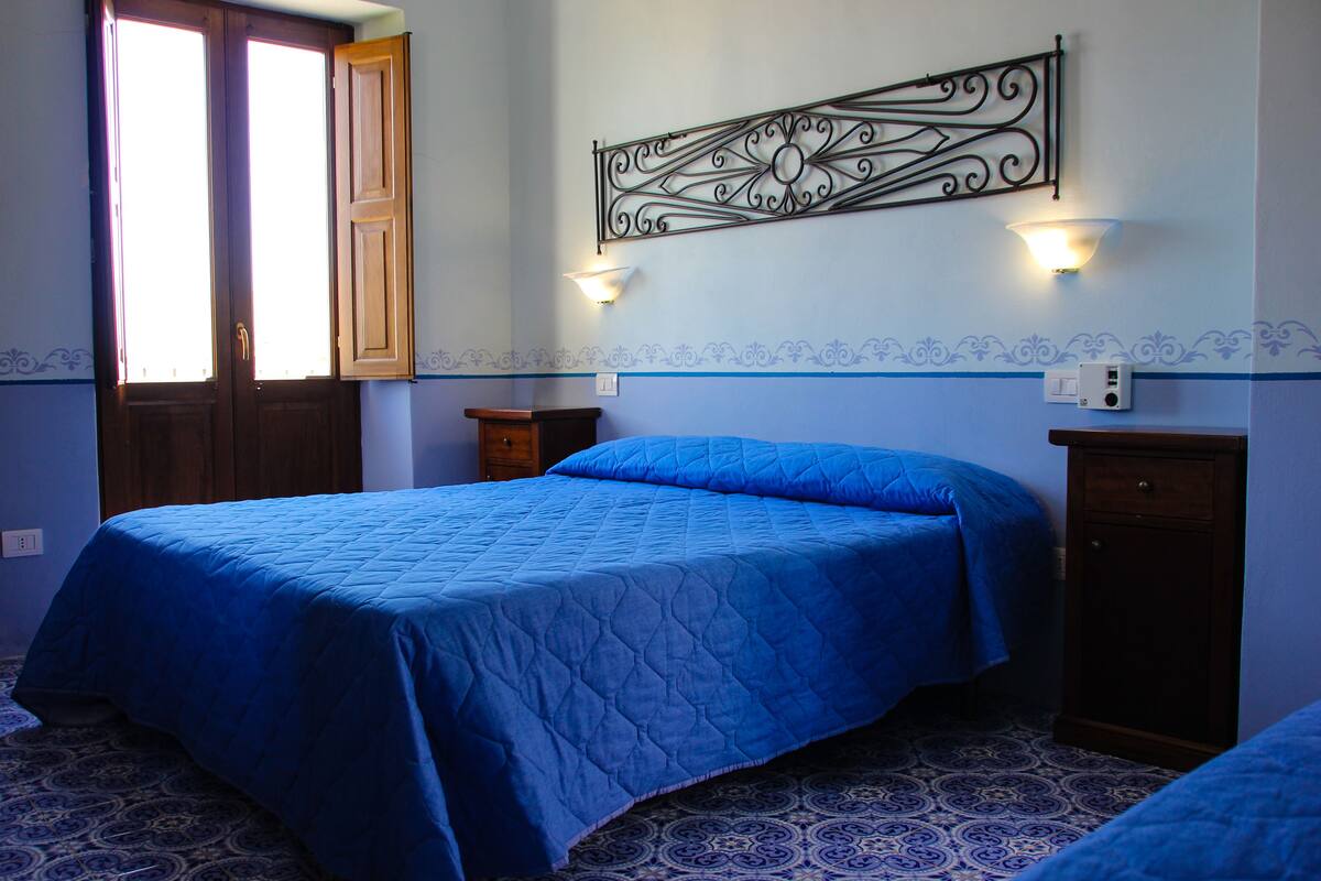A bedroom with a large bed, two bedside tables, a wall-mounted light fixture, and a decorative metal piece above the bed.