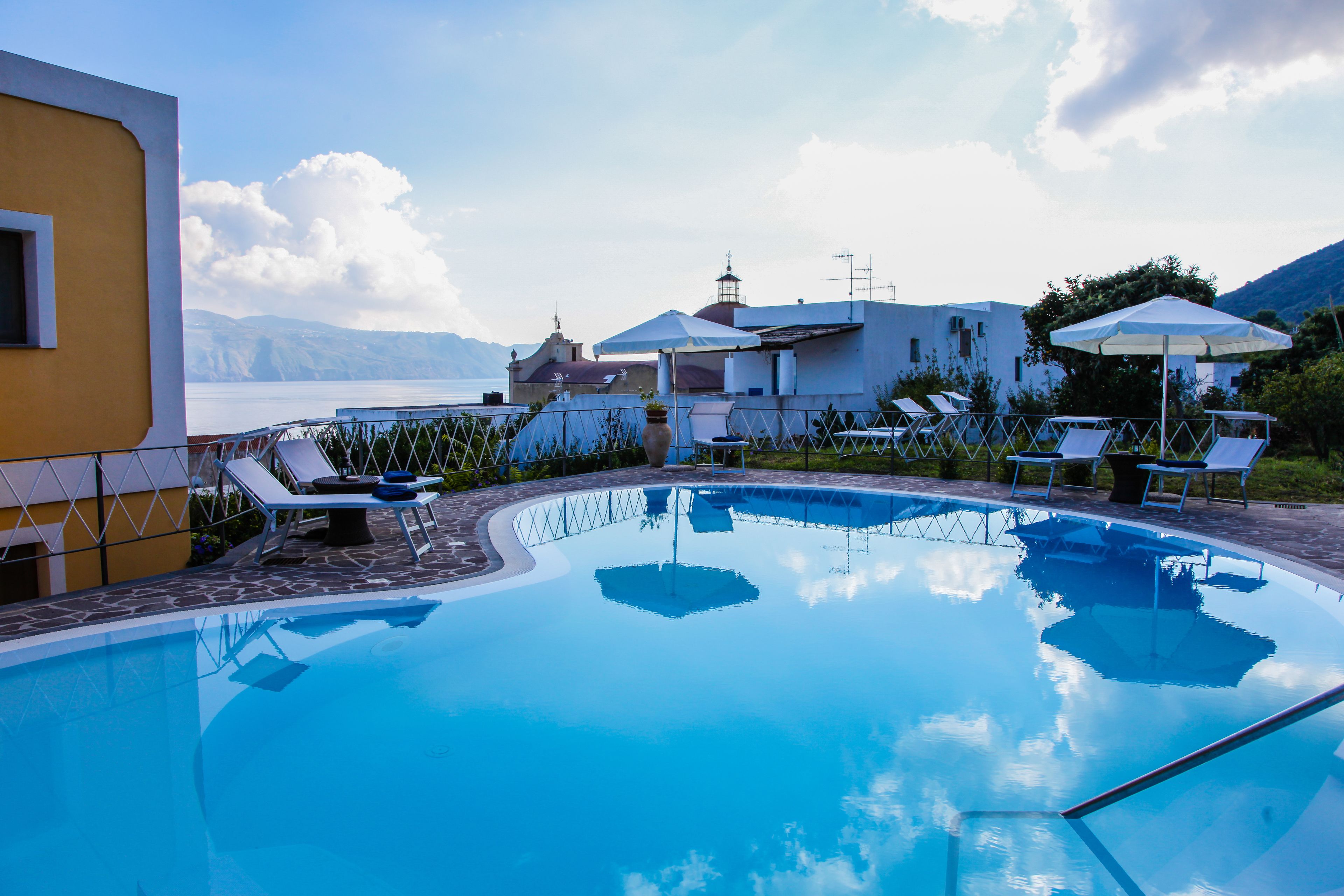 Una piscina con sillas de descanso y sombrillas, rodeada de edificios y vistas al mar.