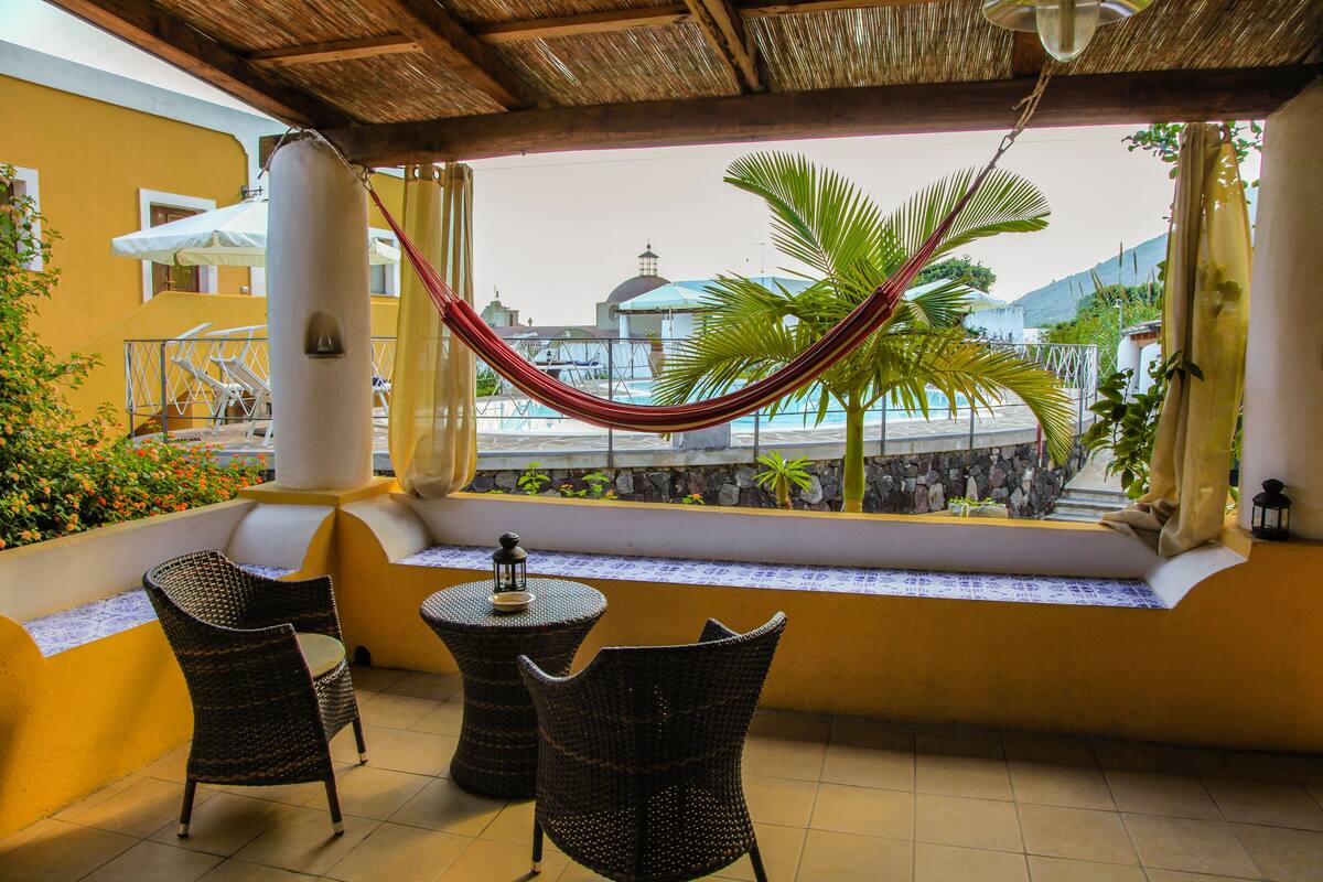 A balcony with wicker chairs and a hammock, overlooking a pool and a building with a dome.