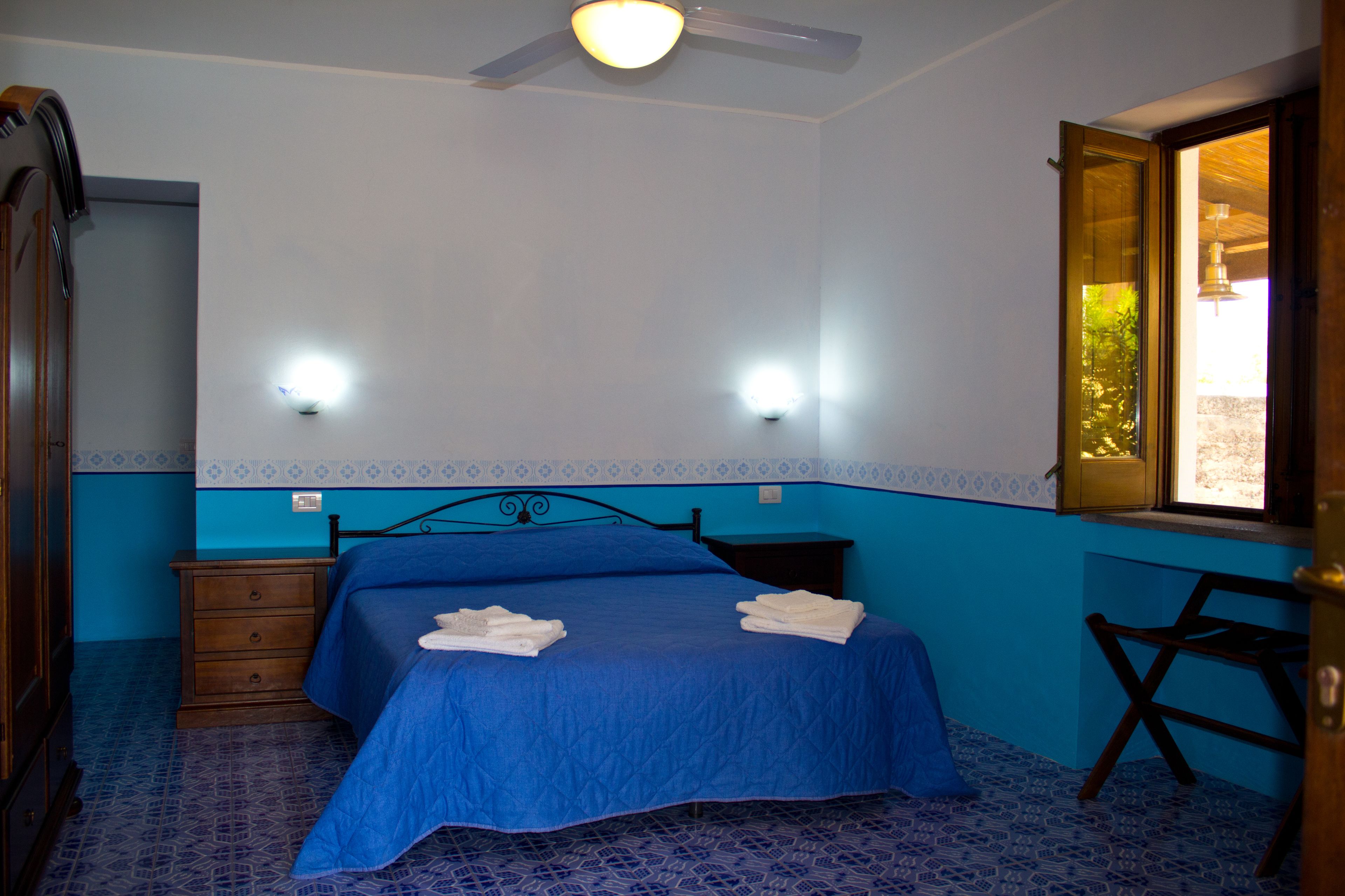 A bedroom with a blue bedspread, two bedside lamps, a wooden nightstand, and a window with a view.
