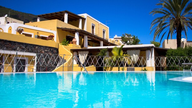 A swimming pool with a clear blue water surface, surrounded by a fence. In the background, there are buildings with yellow and white walls, and a palm tree is visible to the right. - Hotel Arcangelo - Salina (Santa Marina Salina)
