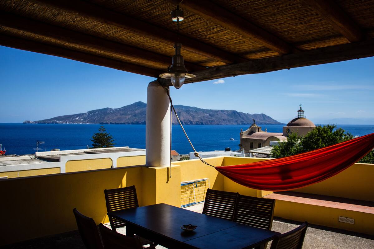 A terrace with a table and chairs, a red hammock, and a view of the sea and mountains.