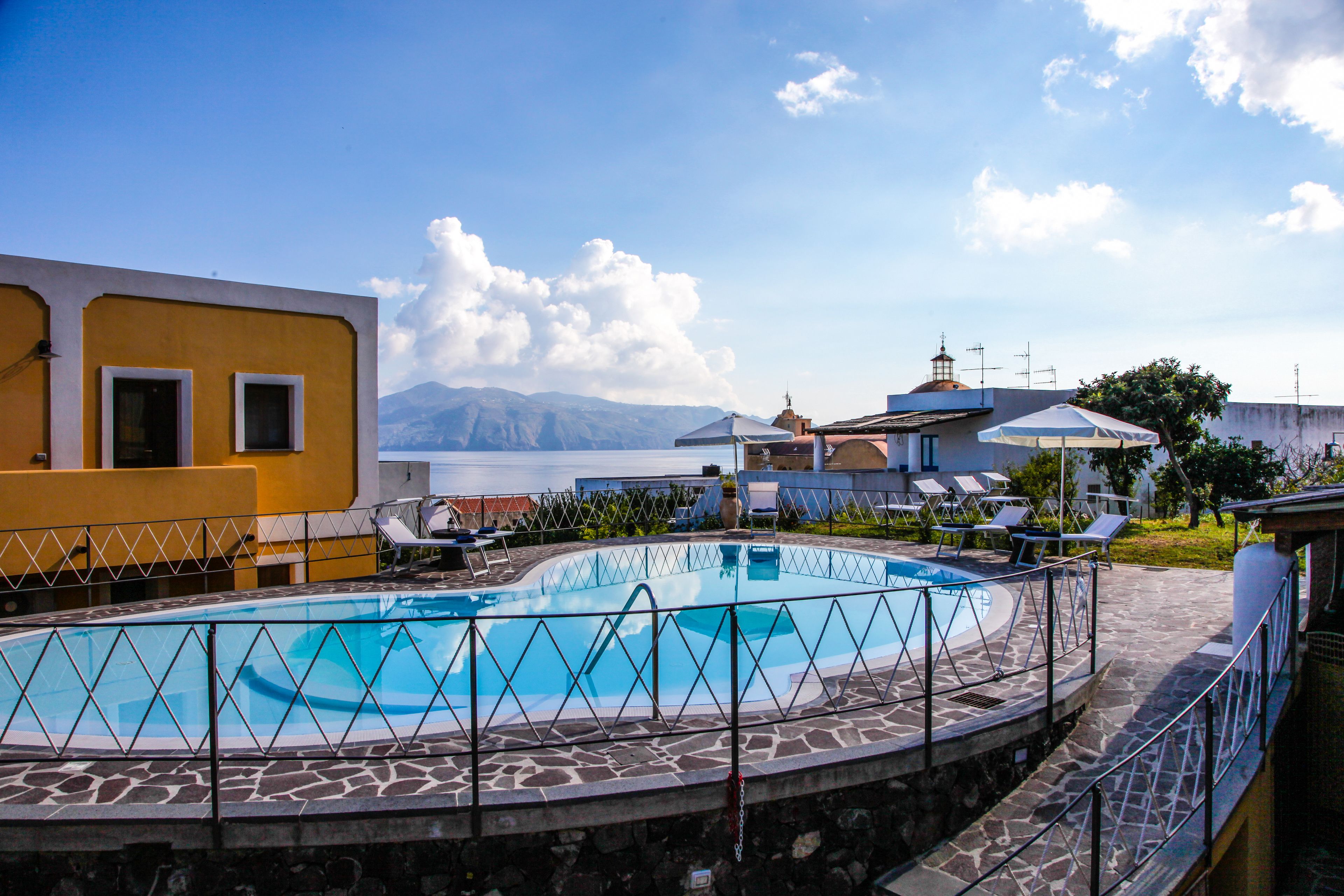 A pool area with a stone deck, lounge chairs, and a view of the sea and mountains.