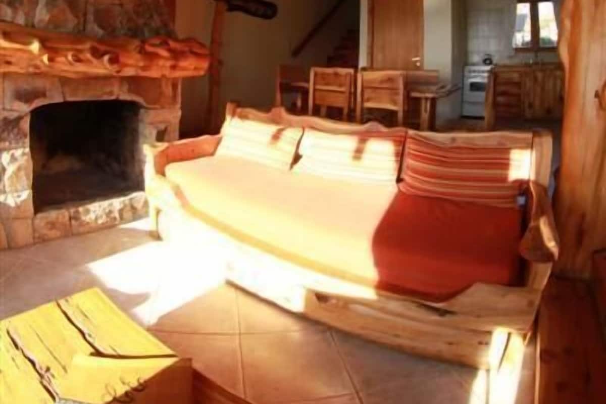 A cozy log cabin interior with a stone fireplace, wooden furniture, and a kitchen area.