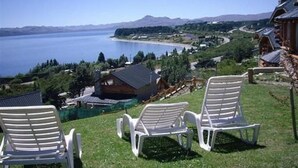 Vista de una propiedad junto al lago con tres sillas de jardín blancas, una cabaña de madera y una vista despejada del lago y las montañas al fondo.