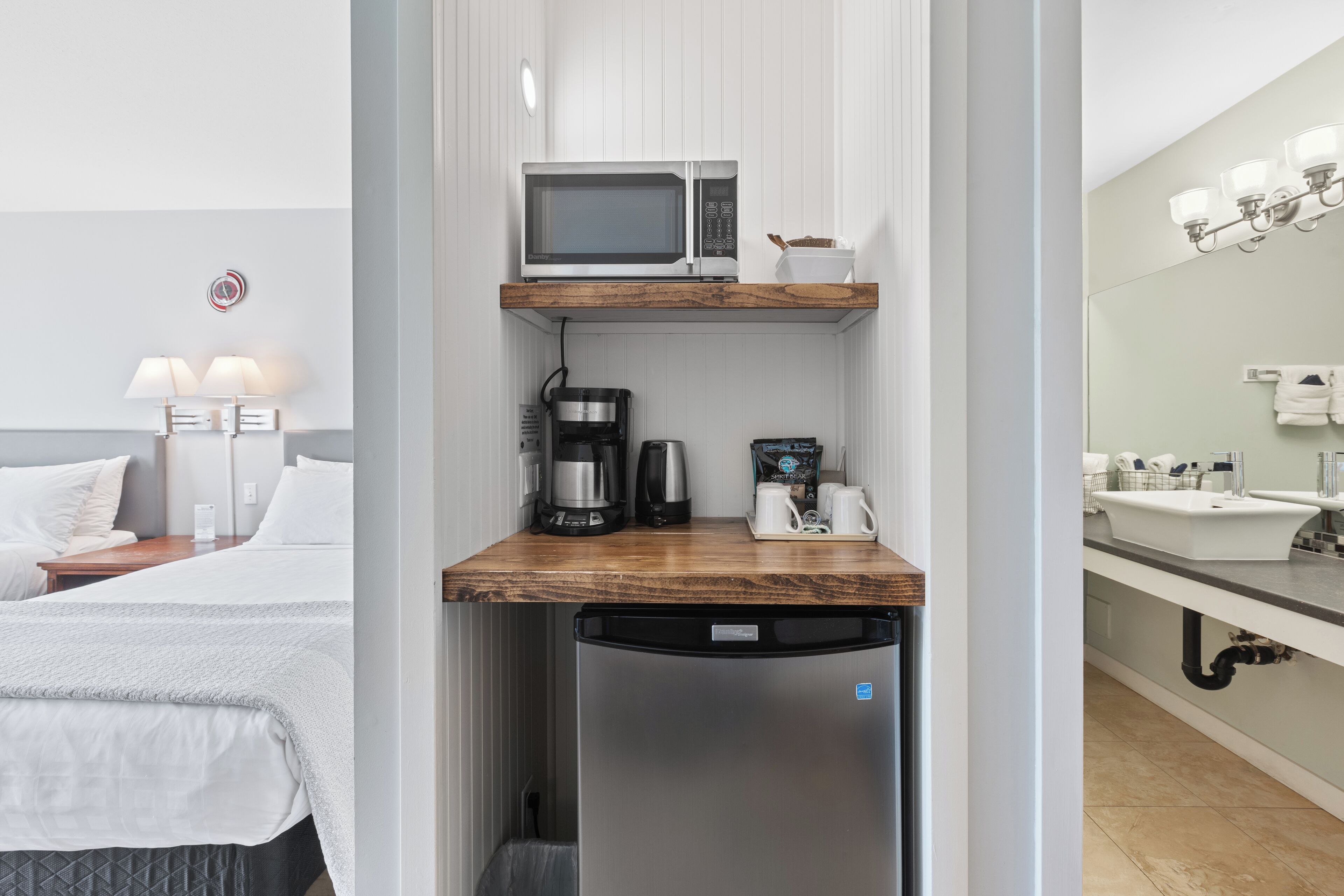 A compact hotel room with a kitchenette, a bed with white linens, a nightstand, and a bathroom with a sink and mirror.