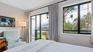 A hotel room with a large bed, a chair, a desk, a lamp, and a window with a view of greenery and mountains. - Tofino Motel Harborview (Tofino)
