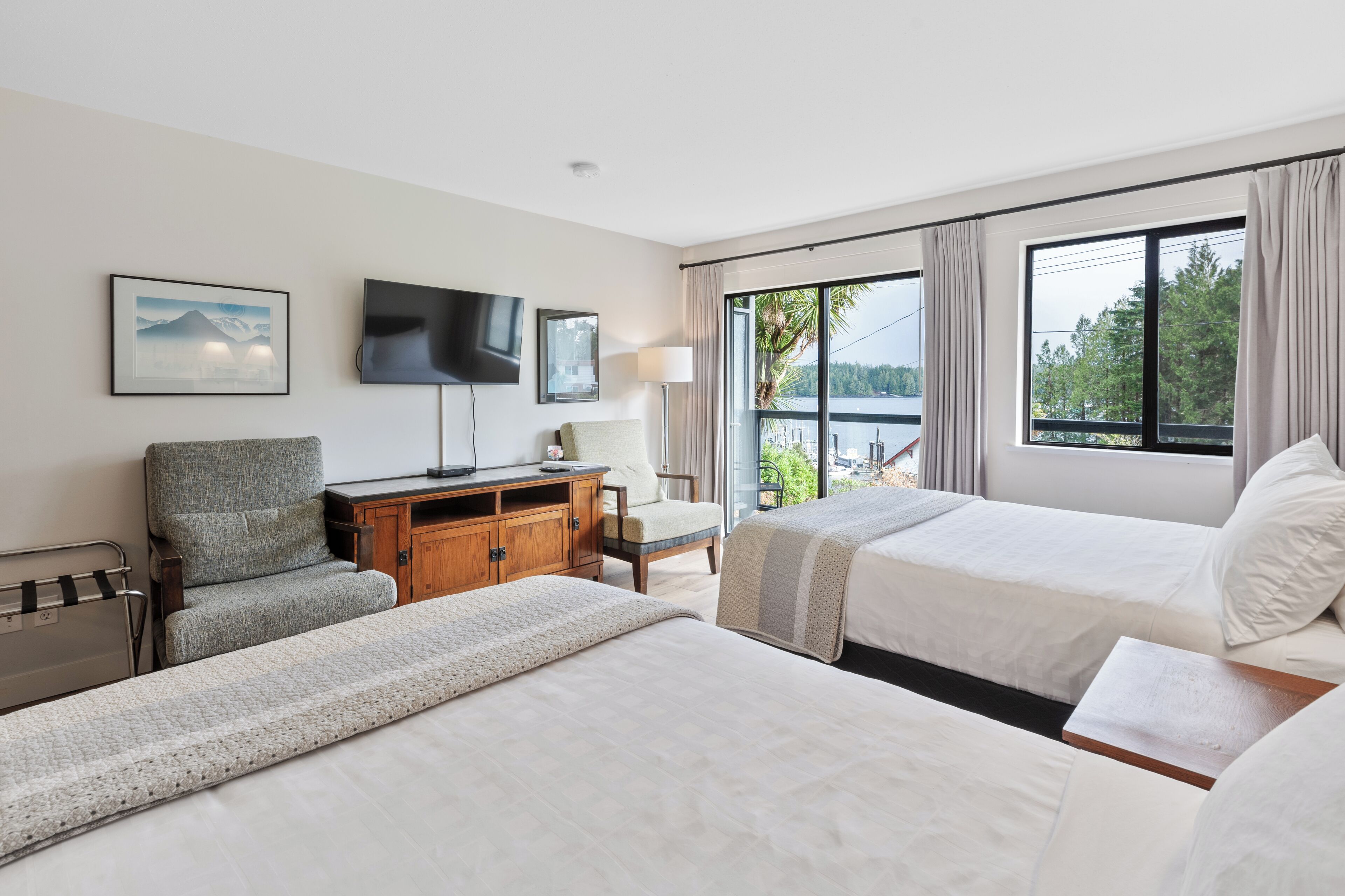 A hotel room with two beds, a desk, a chair, a TV, and a large window with a view of trees and a body of water.