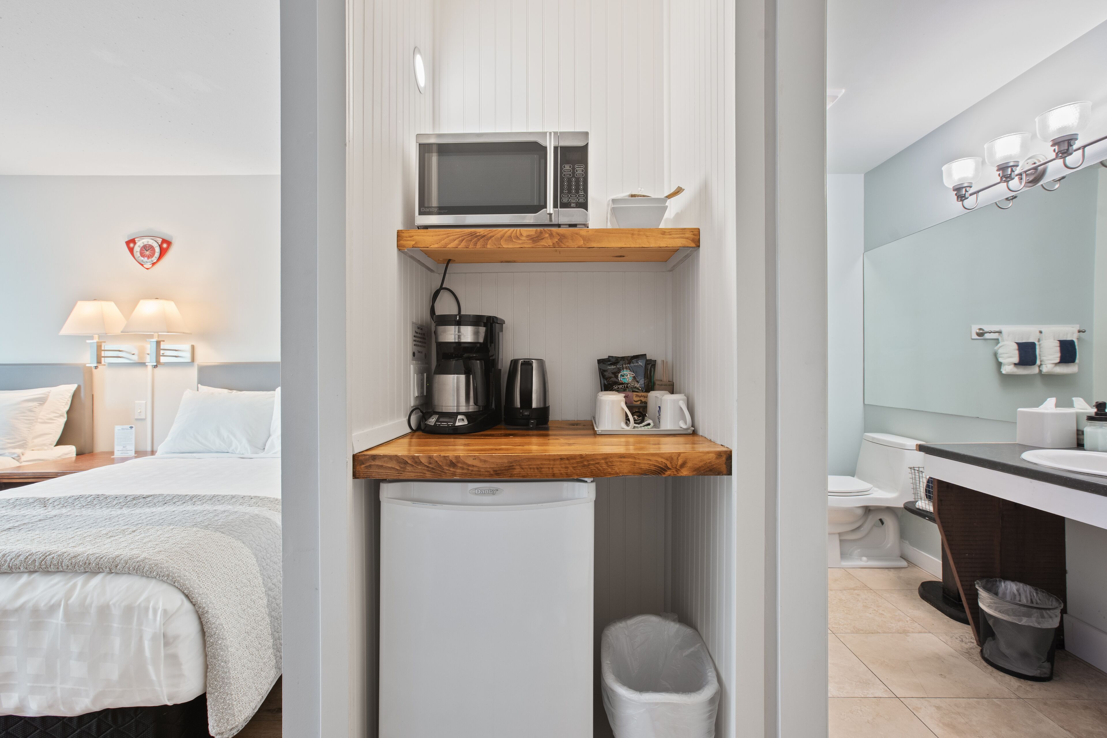 Une chambre d’hôtel compacte comprenant un coin cuisine, un lit, une petite poubelle et une salle de bain avec un lavabo et un miroir.