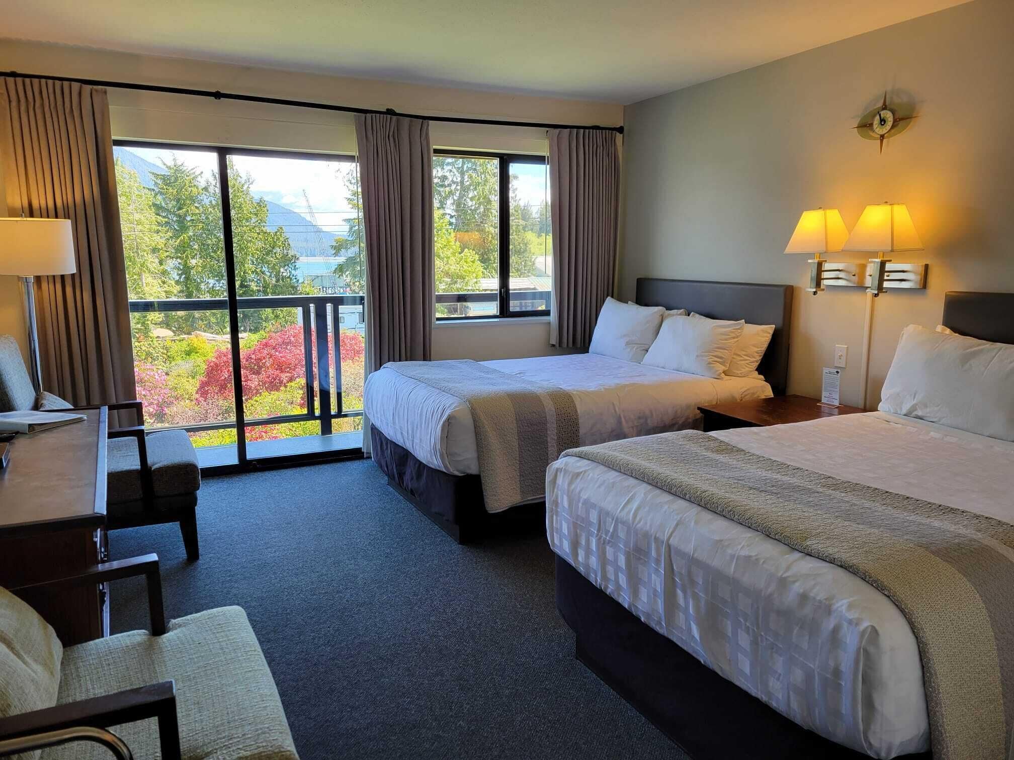 A hotel room with two beds, a desk, and a chair. There is a balcony with a view of trees and mountains.
